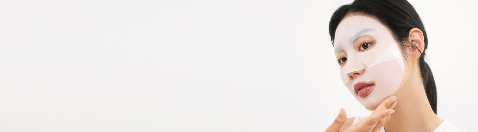 Une femme aux cheveux foncés applique un masque blanc sur son visage, en le pressant délicatement sur sa joue. Le fond est sobre et clair, créant un look minimaliste.