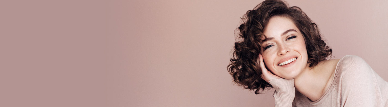 Une femme souriante aux cheveux bruns courts et bouclés et au maquillage léger, vêtue d'un pull beige, pose sa tête sur sa main sur un fond beige doux.