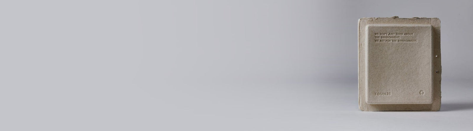 Une boîte fermée, rectangulaire, beige, en carton recyclé avec un design simple, repose sur un fond gris clair uni, positionnée sur le côté droit de l'image.