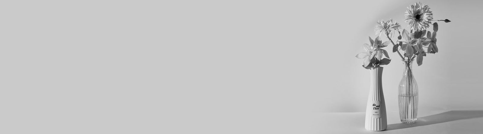Deux vases en verre contenant des fleurs sont placés côte à côte sur un fond gris clair, avec un large espace vide à gauche de l'image. La scène est en noir et blanc.